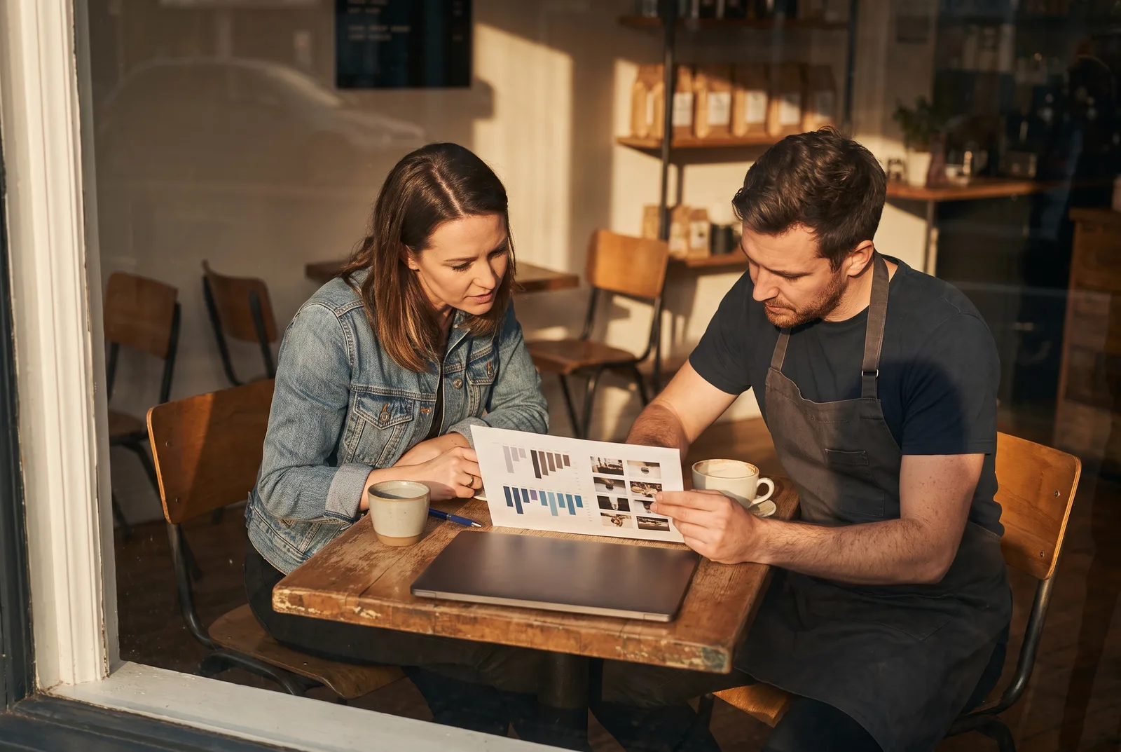 A social media manager and a café owner go through a one-page printed report together at a table, a laptop closed beside them.